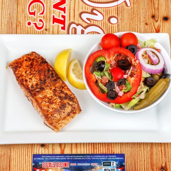Grilled salmon fillet with lemon wedges and a side salad with olives, tomatoes, and red onion.