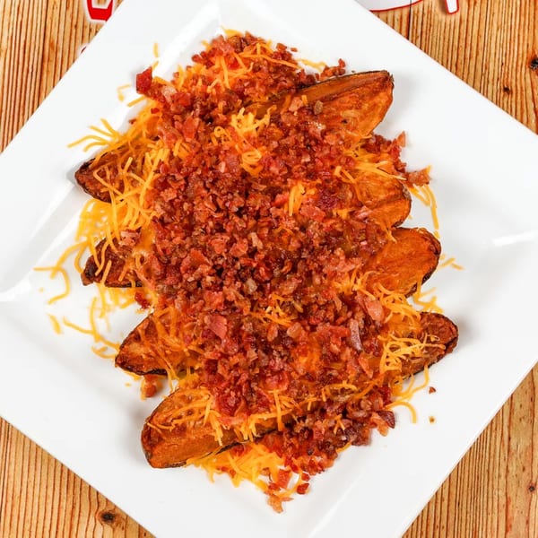 Sweet potato wedges topped with shredded cheddar cheese and crispy bacon bits on a white plate.