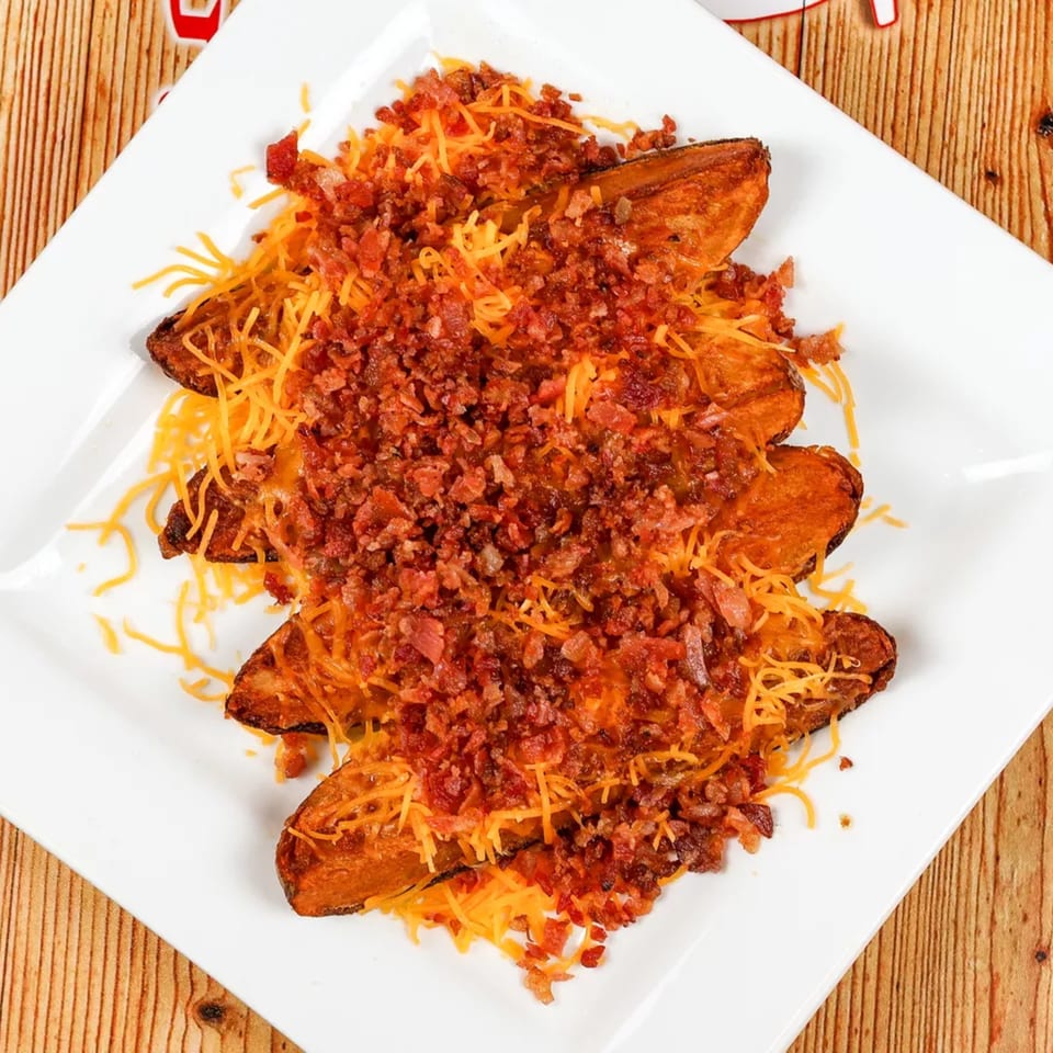 Sweet potato wedges topped with shredded cheddar cheese and crispy bacon bits on a white plate.
