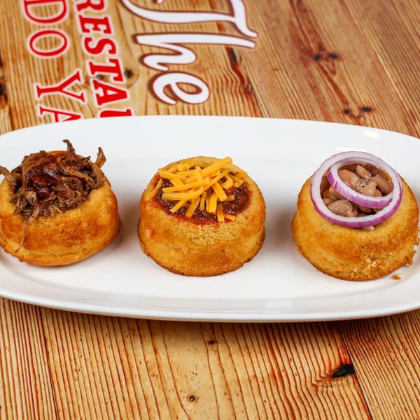 Three cornbread cups filled with pulled pork, chili with cheese, and beans with red onion.