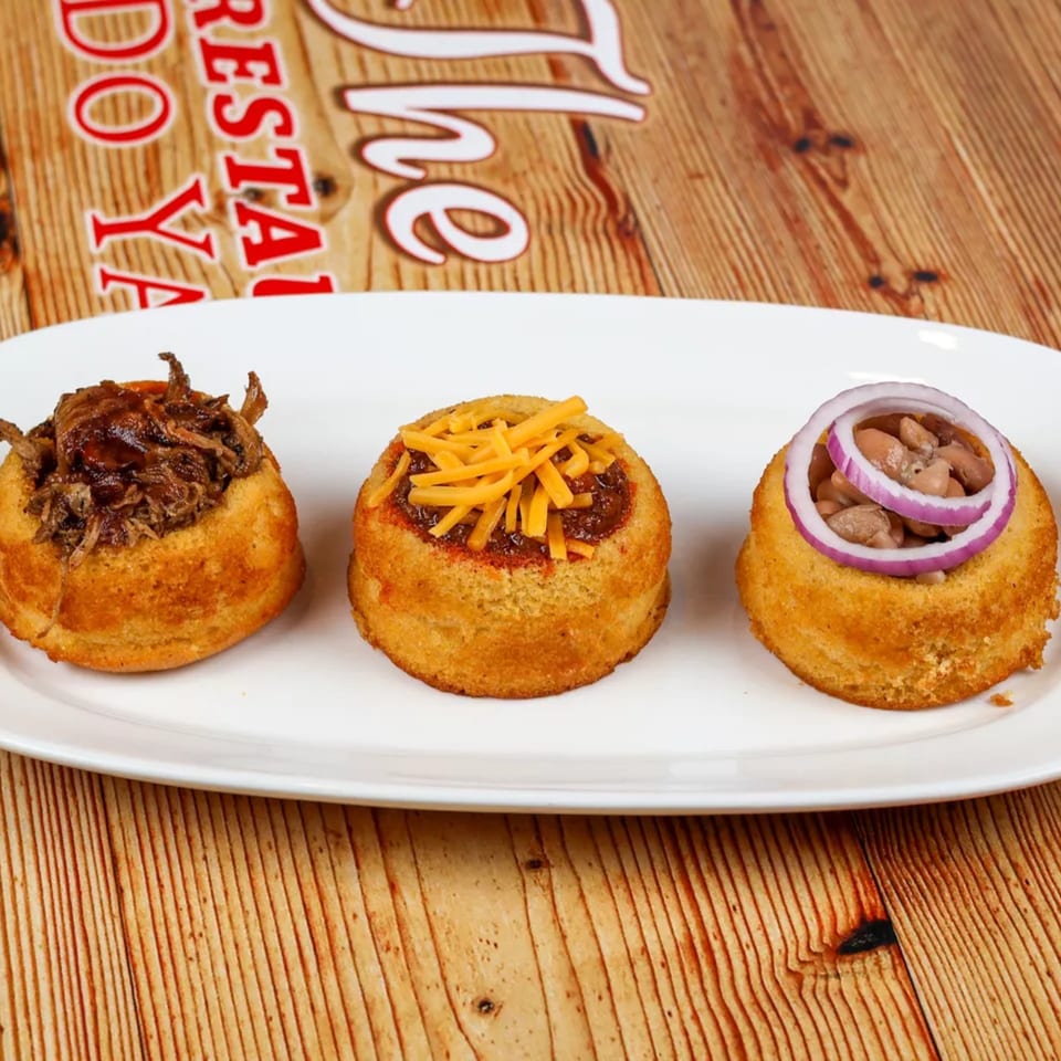 Three cornbread cups filled with pulled pork, chili with cheese, and beans with red onion.