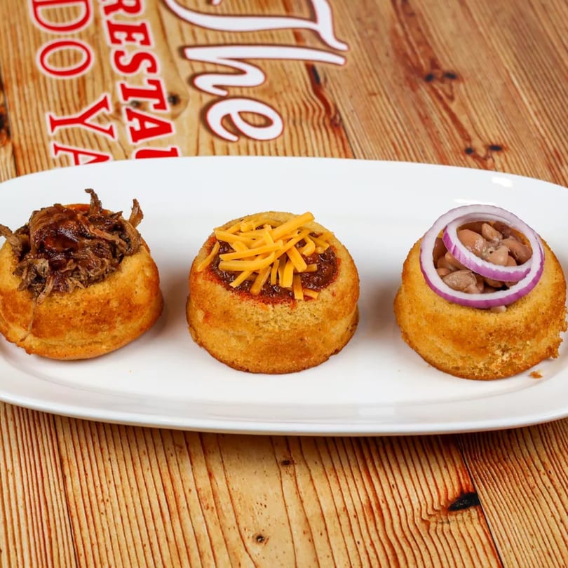 Three cornbread cups filled with pulled pork, chili with cheese, and beans with red onion.